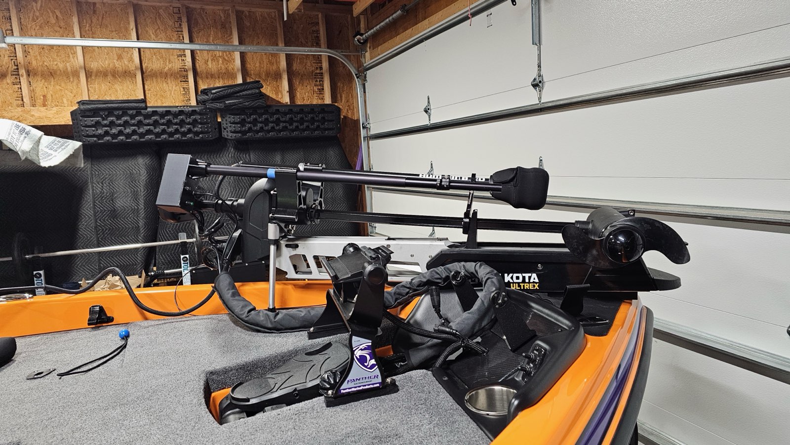 A close-up of a fishing boat's bow in a garage features a Minn Kota Ulterra trolling motor, the Wireless Transducer Pole - First Mate XT (Formerly MotaPole), various fishing electronics, and other equipment on the deck.