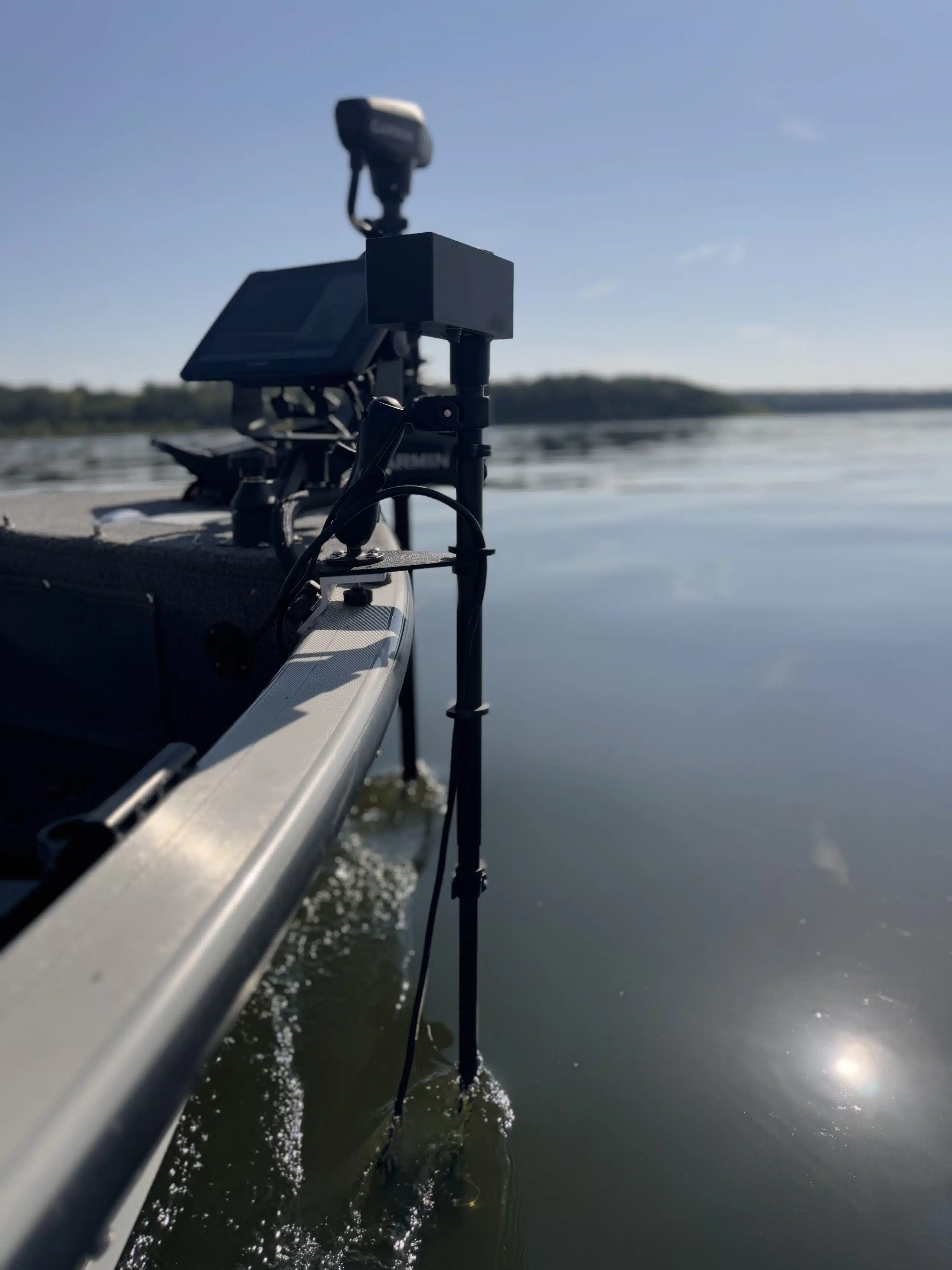 Close-up of a fishing boat using the First Mate XT Wireless Transducer Pole (formerly MotaPole) and an electronic fish finder, gliding on calm water beneath a clear blue sky with distant trees in view.