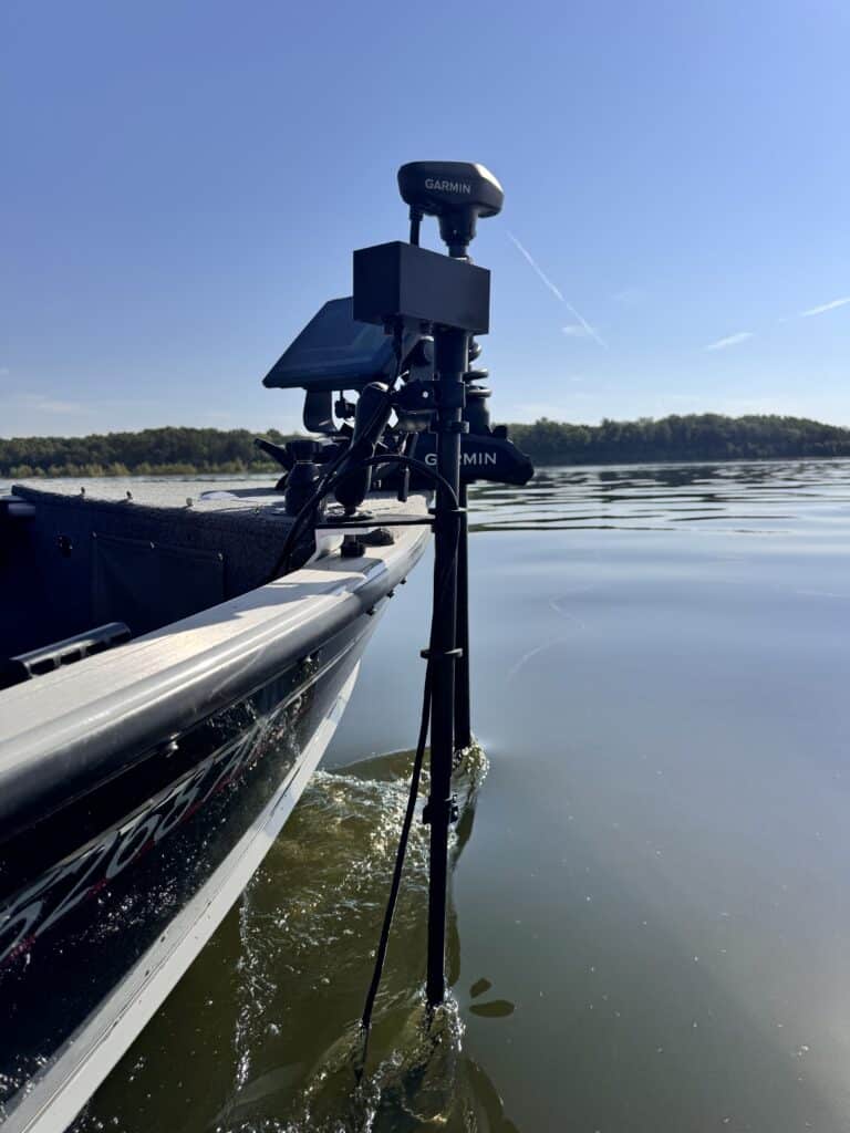 A close-up of a boat on a calm lake shows Garmin electronics and a Wireless Transducer Pole - First Mate XT (Formerly MotaPole), with trees and blue sky in the background.