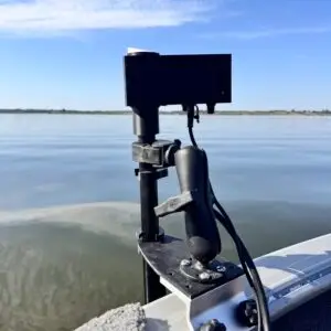 A close-up of an electronic device with wires connected to a Wireless Transducer Pole - First Mate XT (Formerly MotaPole) mounted on a boat's edge, overlooking calm water beneath a blue sky with scattered clouds.