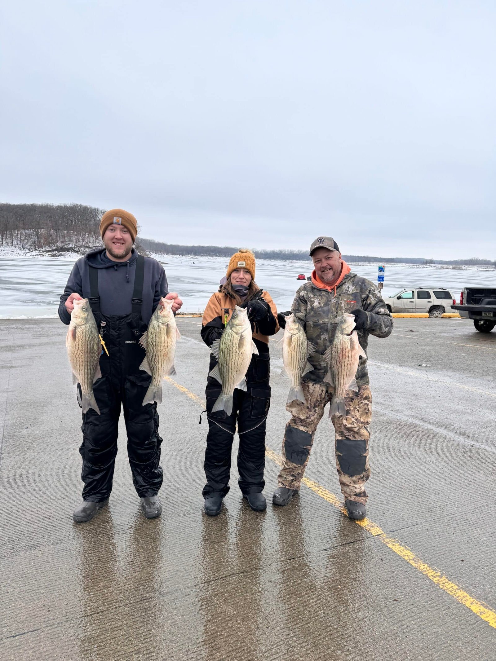 Three people dressed in winter clothing stand on a paved area by a frozen lake, each holding large fish they caught, smiling at the camera. Snow and icy water are visible in the background.
