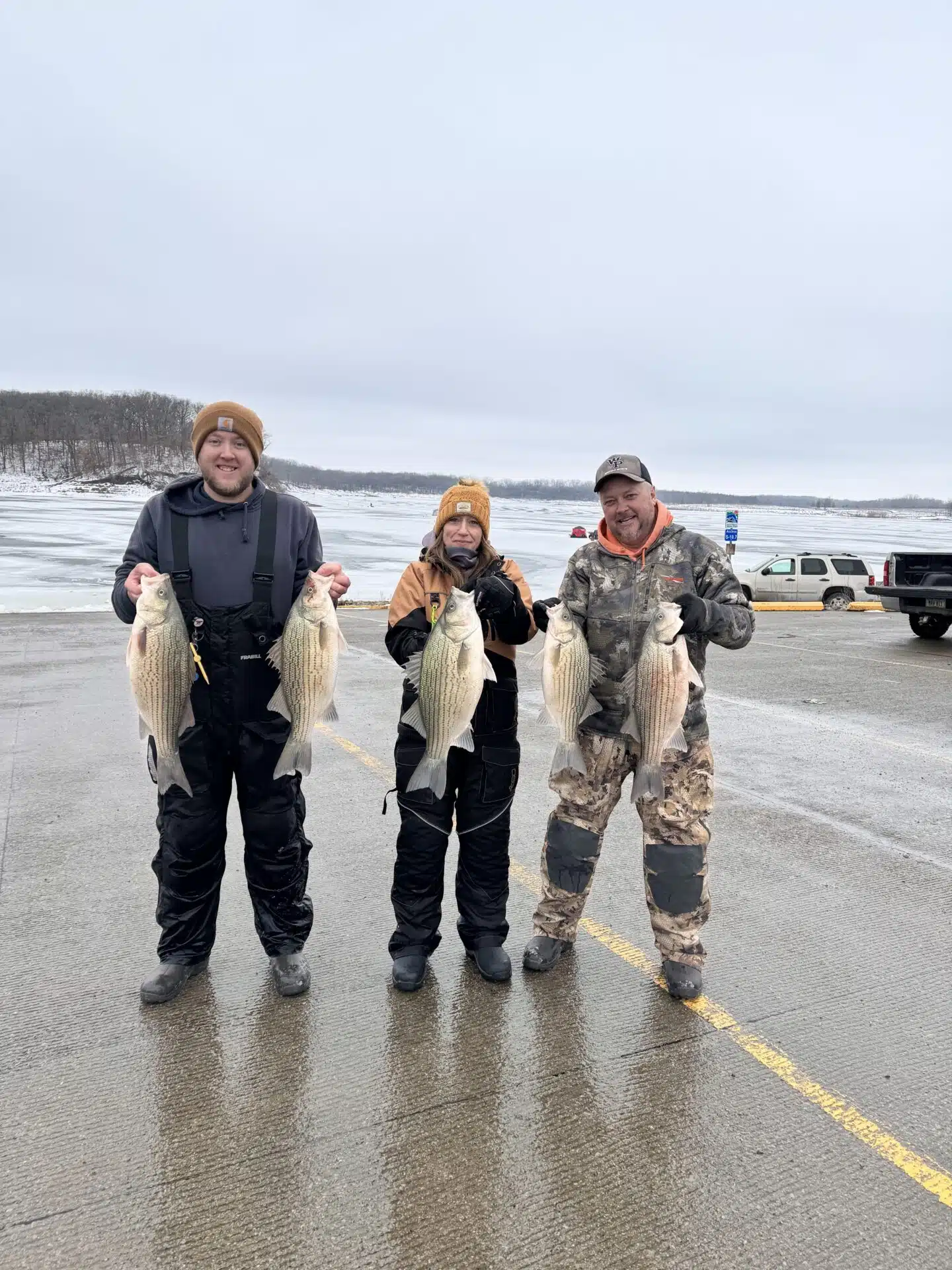 Three people dressed in winter clothing stand on a paved area by a frozen lake, each holding large fish they caught, smiling at the camera. Snow and icy water are visible in the background.