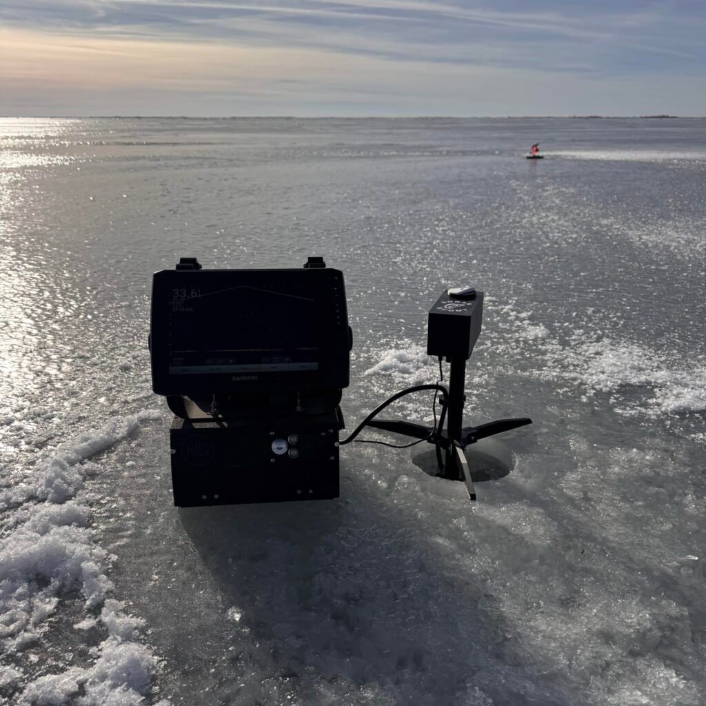A fish finder and sonar device are set up on a frozen lake next to a small hole in the ice, with snowy patches and a distant ice fishing tip-up visible under a cloudy sky.