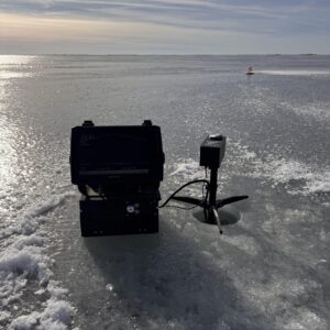 A fish finder and sonar device are set up on a frozen lake next to a small hole in the ice, with snowy patches and a distant ice fishing tip-up visible under a cloudy sky.
