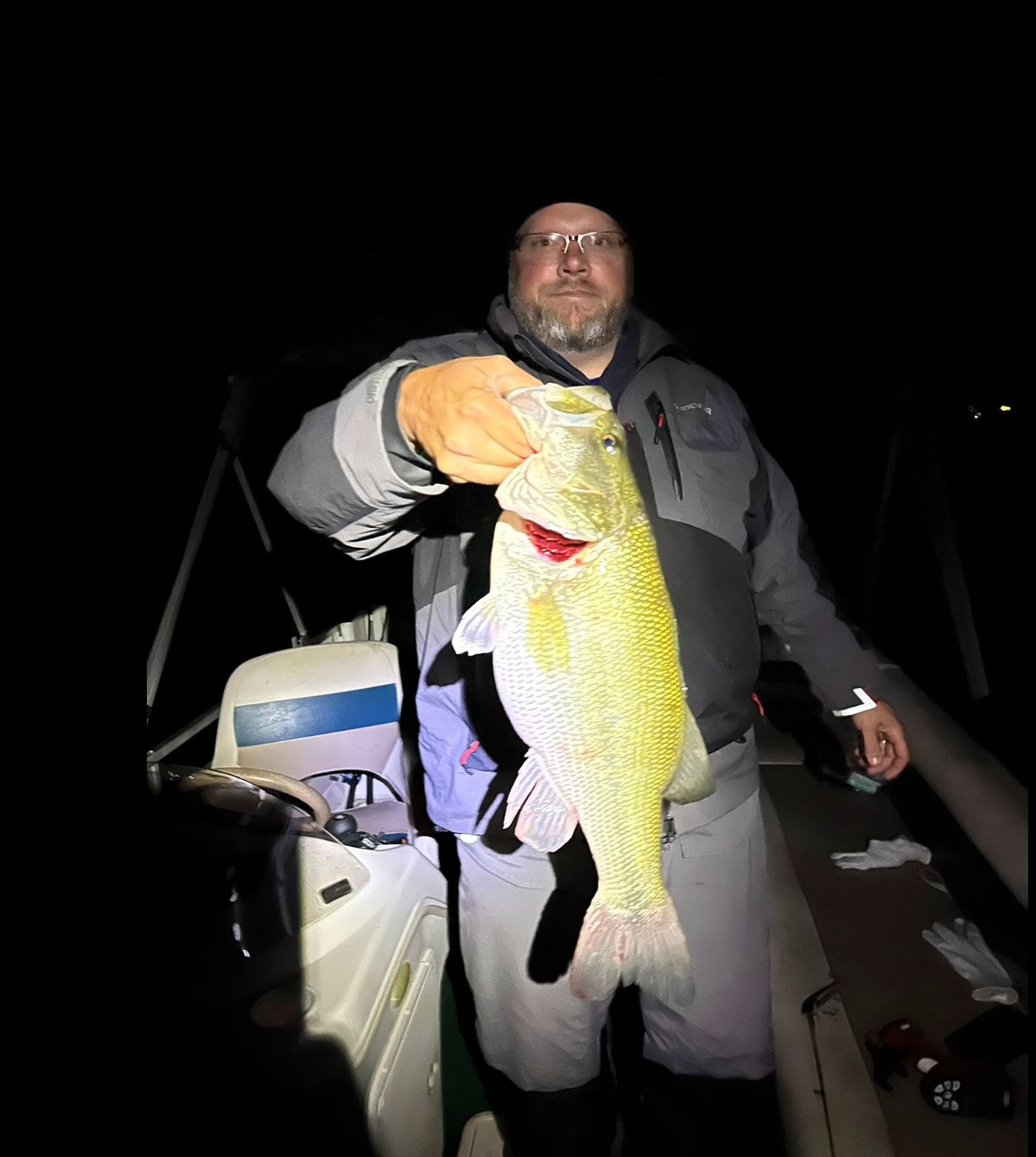 A man wearing glasses and a jacket stands on a boat at night, holding up a large fish by its mouth with his right hand. The fish is illuminated by bright light, highlighting its scales.