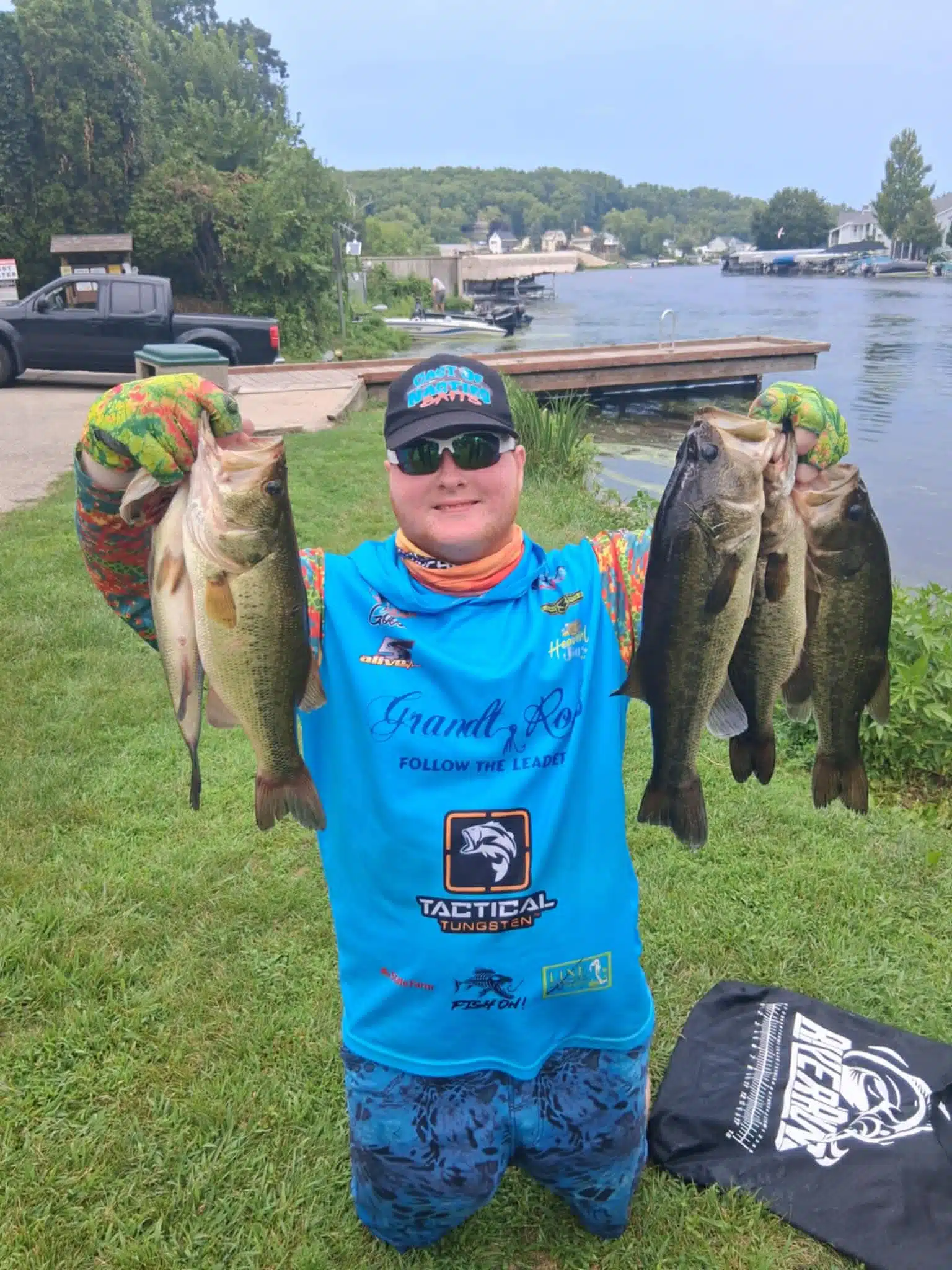 A smiling angler in a blue fishing outfit holds up four large bass, two in each hand, by a lakeside with docks, boats, and trees in the background on a sunny day.