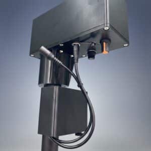A close-up of a black Battery Box mounted on a pole, with multiple cables and connectors attached underneath, set against a neutral gradient background.