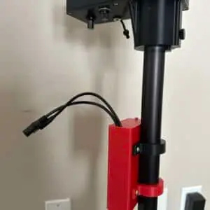 A close-up of a black pole with a red 3D-printed bracket holding two black cables, supporting a Battery Box with buttons and knobs. Electrical outlets are visible on the beige wall in the background.