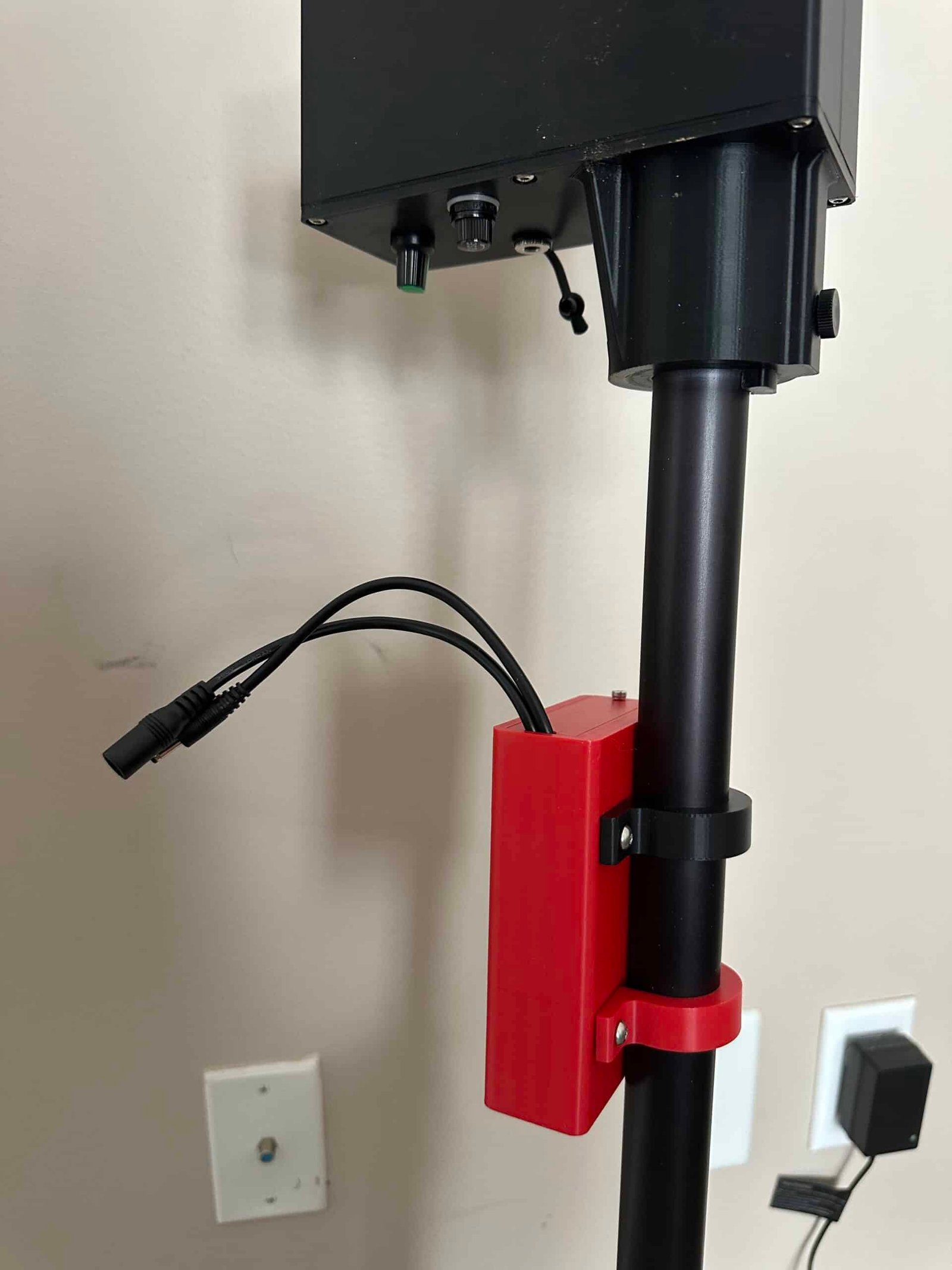 A close-up of a black pole with a red 3D-printed bracket holding two black cables, supporting a Battery Box with buttons and knobs. Electrical outlets are visible on the beige wall in the background.