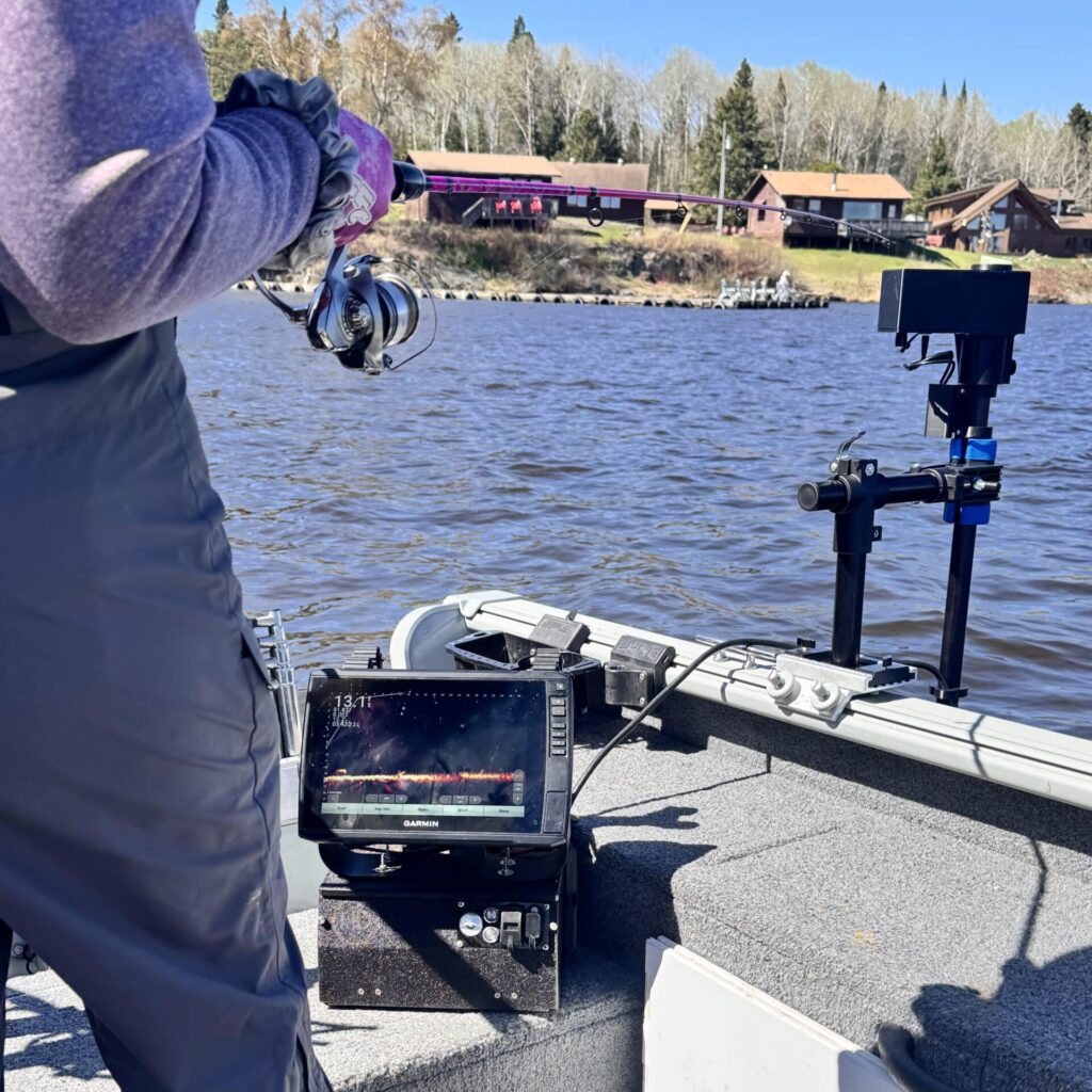 A person stands on a boat using the Universal Transducer Pole Mount with a fish finder, displaying sonar data, as trees and cabins line the far side of the lake under a clear sky.