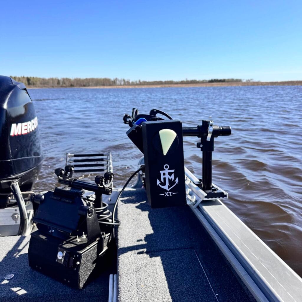 A view from a boat on a lake under a clear blue sky, with fishing gear and a Universal Transducer Pole Mount at the boat’s edge beside a Mercury outboard motor. Forested shoreline is visible in the background.