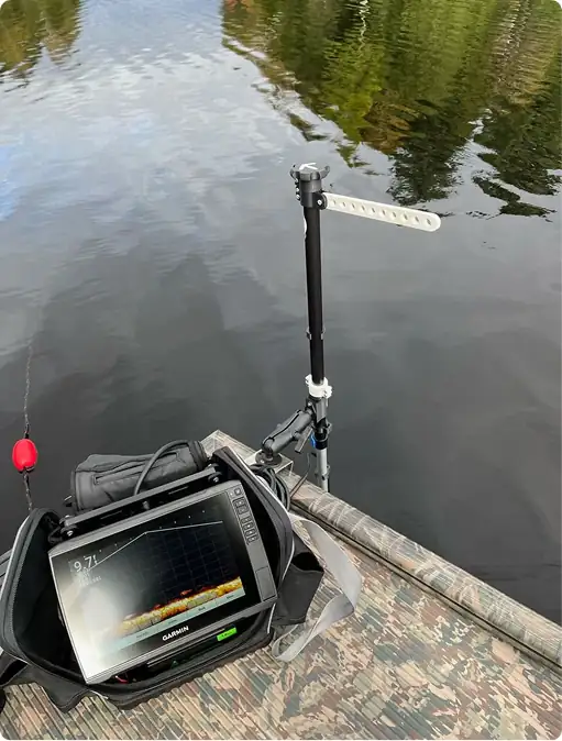 A fish finder device with a screen showing sonar readings is set up on a dock above calm water, with a rod and sensor mounted at the edge, surrounded by reflections of trees.