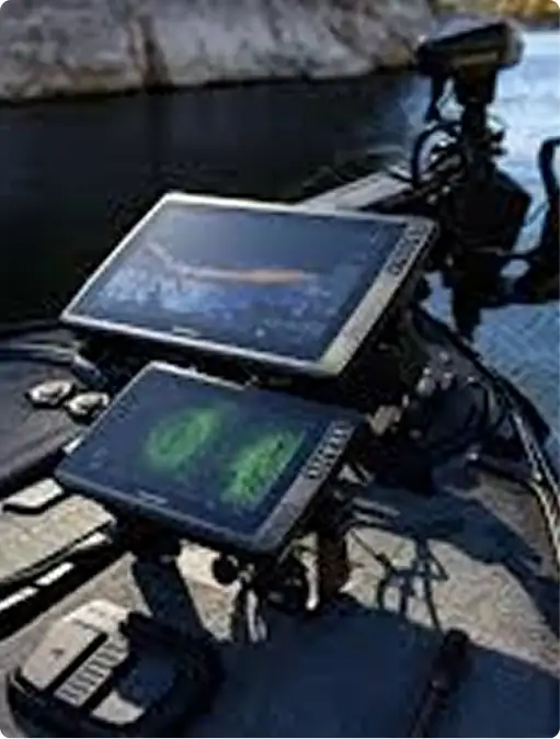 Two electronic fish finders with display screens are mounted on a boat, showing digital sonar and mapping data, with water and rocky shoreline visible in the background.