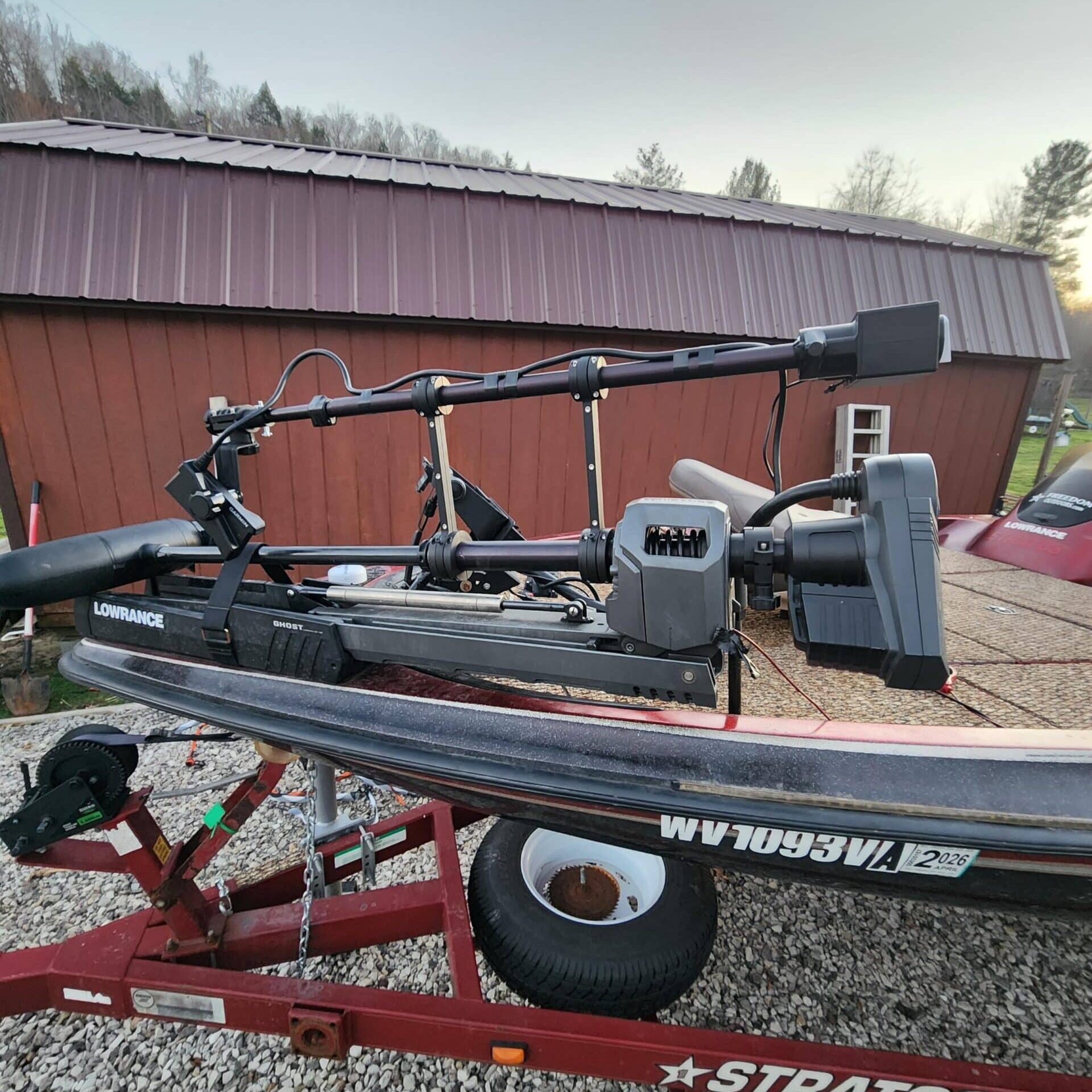 A close-up view of a bass boat on a red trailer, featuring mounted electronic fish finders and trolling motors, parked on gravel beside a brown building with a tin roof.