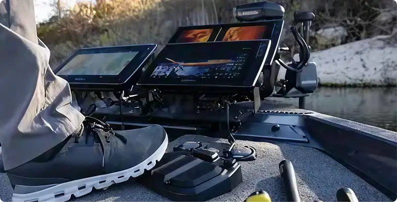 A person stands on a boat deck next to two large fishfinder screens and various fishing equipment, with a riverbank and trees visible in the background.
