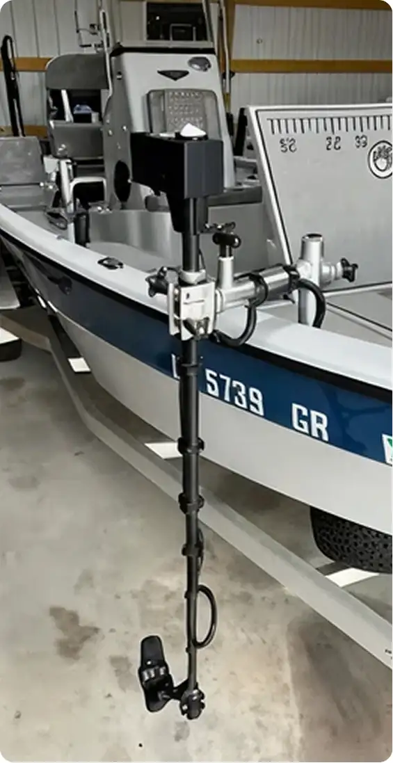 A close-up view of a black trolling motor mounted on the side of a white and blue fishing boat, which is parked indoors on a concrete floor. A measuring board is visible on the boat’s deck in the background.