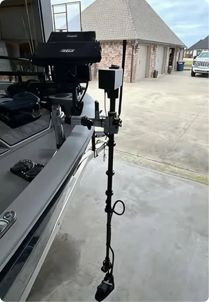 A metal pole with mounted electronic equipment is attached to the front of a boat parked in a driveway near a brick house with garages and a white truck in the background.