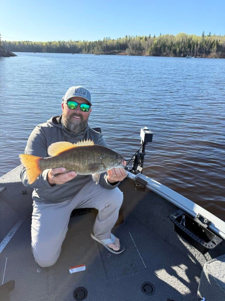 A man in sunglasses and a cap kneels on a boat holding a large fish with orange fins, with a calm lake and tree-lined shore in the background on a sunny day.