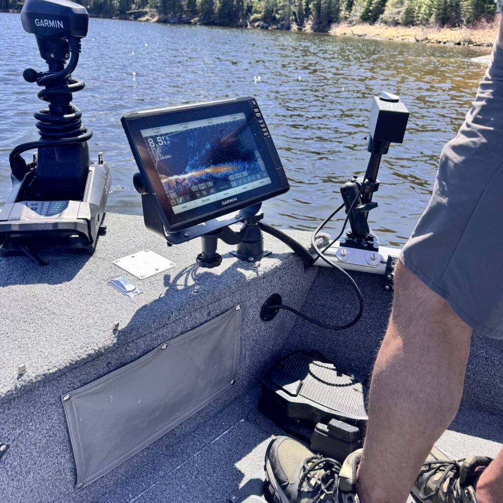 A person stands on a fishing boat equipped with sonar and navigation devices, including a Garmin unit and a mounted screen displaying sonar data, on a calm lake surrounded by trees.