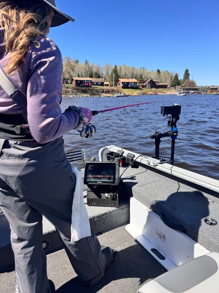 A person in fishing gear stands on a boat holding a fishing rod over a lake, with electronic equipment on the boat and cabins and trees visible along the shoreline under a clear blue sky.