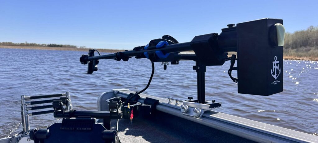 A close-up of a mounted trolling motor and electronic equipment on the deck of a fishing boat, with calm water and a clear blue sky in the background.