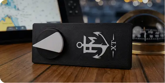 A black rectangular device with a large silver knob and a white anchor symbol on top, sitting on a wooden surface near nautical instruments and a digital map display.