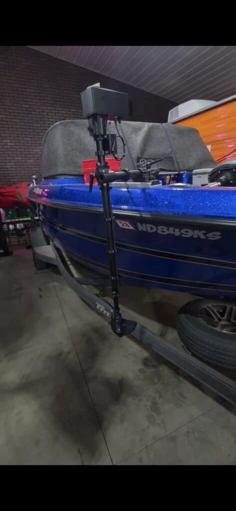 A blue fishing boat on a trailer inside a garage, with a mounted black pole and electronic device attached to the boats side. The boat is parked on a concrete floor near a brick wall.
