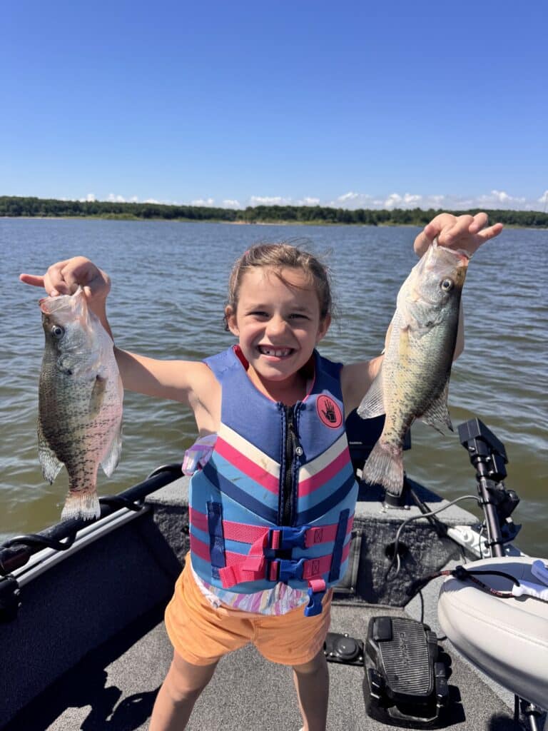 Young girl in a life jacket holding up two crappie on a boat
