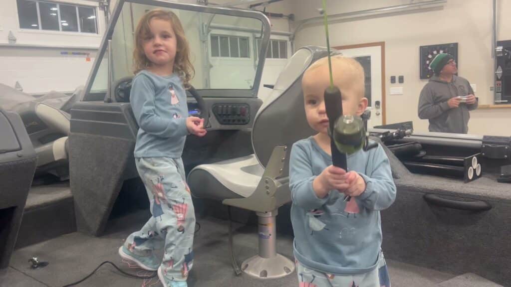 Two grandkids playing in the boat in the First Mate shop, one at the wheel and one holding a rod